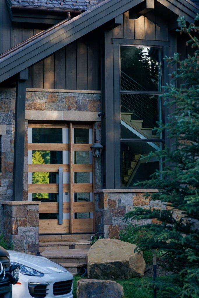 Entrance of a modern rustic house with wooden door and glass window featuring outdoor green plants.